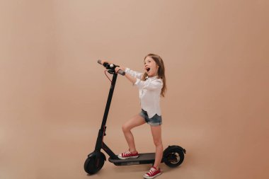 Beyaz gömlekli, şortlu, heyecanlı küçük bir kızın bej arka planda elektro motorsikletle oynayıp eğlendiği uzun bir fotoğraf.. 
