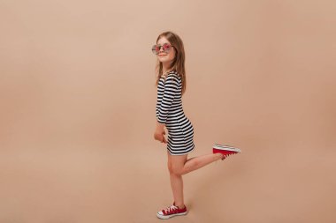 Güzel elbiseli ve gözlüklü küçük kızın mutlu bir gülümsemeyle kameraya poz verip bacağını kaldırdığı uzun bir fotoğraf. .