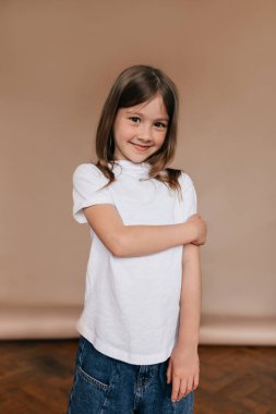 Beyaz tişörtlü, kot pantolonlu, bej arka planda duran sevimli şık çocuk. Küçük esmer kızın eğlendiği bir stüdyo fotoğrafı..