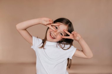 Mutlu, heyecanlı, mutlu duyguları olan küçük bir kız bej arka planda mutluluk belirtileri gösteriyor ve gülümsüyor. 