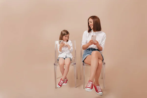 全长照片中的年轻女子穿着白衬衫和短裤 坐在椅子上 头戴米黄色背景的智能手机 看着她的小女儿 小女孩和妈妈一起玩智能手机 — 图库照片