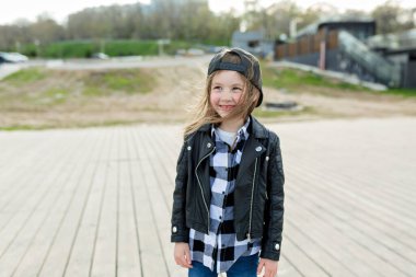 Sevimli sevimli kız otel kareli gömleği ve deri ceketli otel şapkası ahşap iskelede duruyor ve ılık bahar gününde gülümsüyor. 