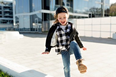 Heyecanlı küçük kız kot pantolon, kareli gömlek ve deri ceket giyiyor. Dışarıda eğleniyor ve şehrin modern binalarına karşı sıcak bir otel düzenliyor. 