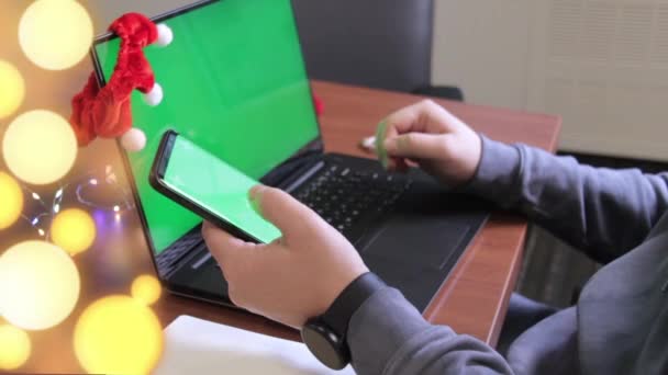 Vista lateral de un freelancers manos escribiendo en el teléfono inteligente con pantalla verde junto a la computadora portátil. Hombre escribiendo en el teléfono móvil. Espíritu navideño. Vista de cerca, oficina. maqueta — Vídeos de Stock