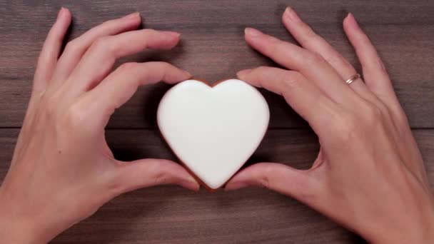 Τα γυναικεία χέρια κρατούν λευκό μπισκότο μεγάλης καρδιάς στο κέντρο και το σηκώνουν πάνω σε ξύλινο φόντο. Έννοια αγάπης. Ημέρα του Αγίου Βαλεντίνου. Αντιγραφή χώρου — Αρχείο Βίντεο