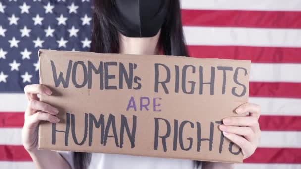 黒いフェイスマスクの若い女性は 女性の権利を持つ段ボールを保持している人権は背景にアメリカの国旗に署名しています 反中絶法に抗議する少女 フェミニストの力 独立した自由 — ストック動画