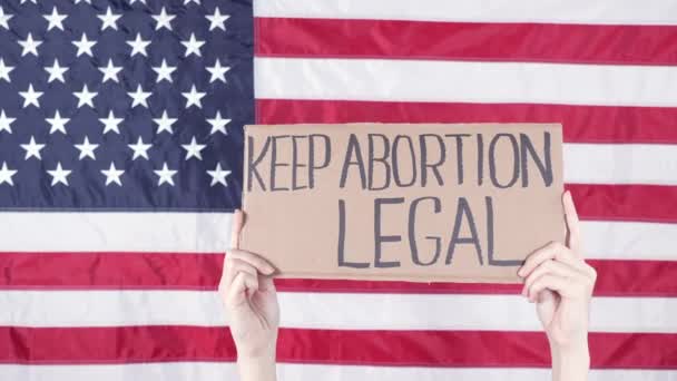 標識を持っている女性は 背景に中絶法的アメリカの旗を保ちます 反中絶法に対する抗議 女性のストライキ 女性の権利の自由 — ストック動画