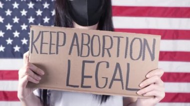 Siyah maskeli genç bir kadın arka planda Amerikan bayrağına karşı Kürtaj Hukukunu Koru tabelası olan bir mukavva taşıyor. Kürtaj karşıtı yasaları protesto eden bir kız. Feminist güç. Kadın Hakları Özgürlüğü