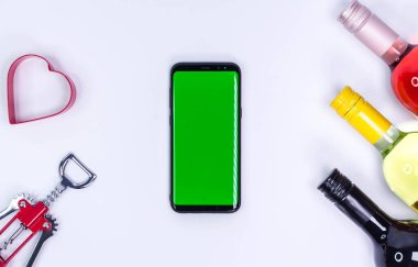 Kırmızı, gül ve beyaz şarap şişeleri tirbuşonlu ve kalbi beyaz masada. Model telefon. Yeşil ekran. Kopyalama alanı olan üst görünüm. Mesaj için yer var. Şarap şişesi boyunları.