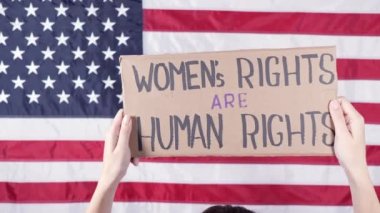 Woman from the back holding a sign Womens Rights Are Human Rights US flag on background. Protest against anti abortion law. Womens strike. Womens rights freedom