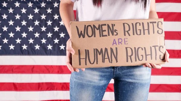 Young woman protester holds cardboard with Womens Rights Are Human Rights sign against USA flag on background. Feminist power. Equal opportunity Womens rights freedom