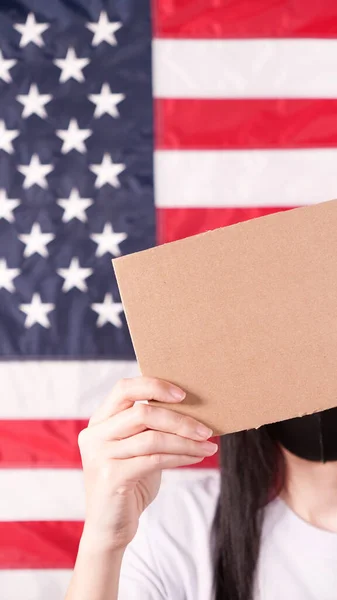 Unrecognizable woman showing empty cardboard banner. Hands holding blank poster from a cardboard box. Space for your slogan, logo or advertisement. Banner design concept. American flag on background.