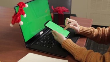 Bilgisayarın yanında yeşil ekranlı akıllı telefondan yazı yazan serbest çalışanların yan görüntüsü. Cep telefonunda yazan bir kadın. Noel tatili ruhu. Yakın çekim, ofis. maketleme.