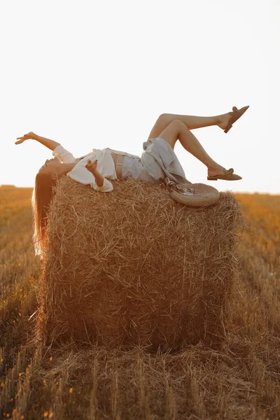 Eine junge Frau mit langen Haaren, Jeansrock und hellem Hemd liegt im Sommer bei Sonnenuntergang auf einem Strohballen im Feld. Frauenporträt in natürlicher ländlicher Umgebung. Ökotourismus-Konzept. — Stockfoto