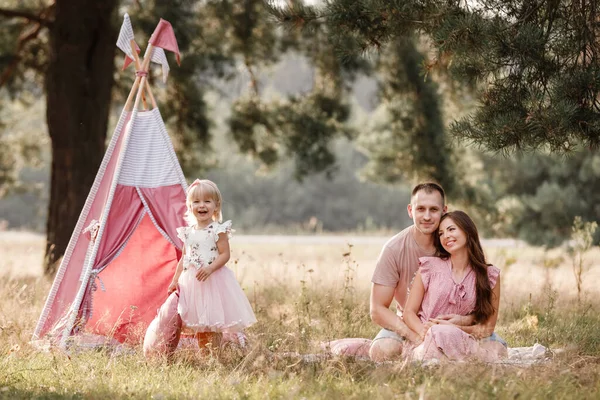 Maman, papa et petite fille sont assis à côté de la décoration wigwam dans le parc. Famille passer du temps en plein air en été, s'amuser ensemble. Fille sont habillés en robe rose — Photo