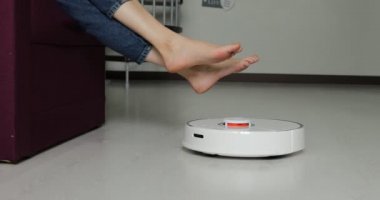 Robot elektrikli süpürge evde çıplak ayaklı bir kadın geziyor. Akıllı bir elektrikli süpürge zemini temizlemek için geçtiğinde kadın ayaklarını kaldırır. Modern hayatta hijyen, ev aletleri ve robotlar kavramı.