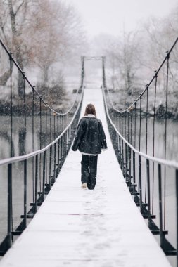 Bir kadın kış günü asma köprüde nehrin üzerinde yürüyor. Soğuk karlı bir günde, kalın elbiseli genç bir kız tahta bir yaya köprüsünde duruyor..
