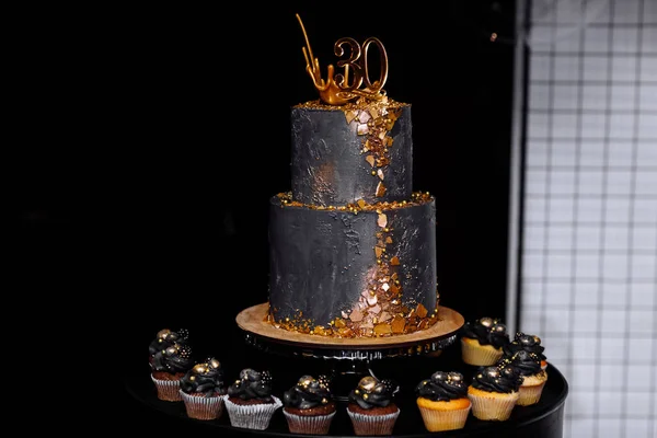 festive two-tier black cake with gold decoration for the 30th anniversary. many chocolate and vanilla cupcakes with luxury decoration. candybar for birthday.