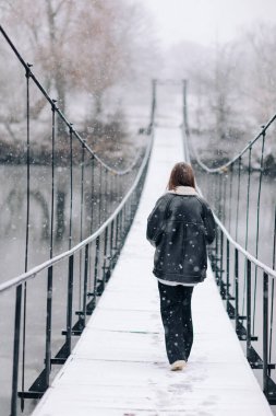 Bir kadın kış günü asma köprüde nehrin üzerinde yürüyor. Soğuk karlı bir günde, kalın giysili genç kız tahta bir yaya köprüsünde duruyor. arka plan.
