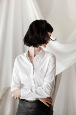 Kollarını kavuşturmuş, kameraya gülümseyen ve eğlenen arkadaş canlısı, kendine güvenen genç bir kadın. Beyaz gömlekli ve deri etekli kız stüdyoda oturuyor..