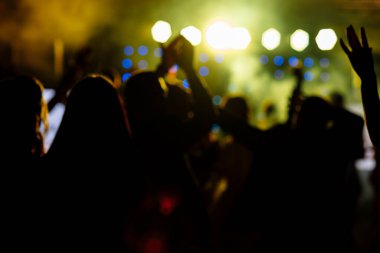 Parlak sarı sahne ışıkları önünde tezahürat yapan kalabalık. Disko gece kulübünde dans eden ya da müzik festivalinde konser veren insanların silüeti.