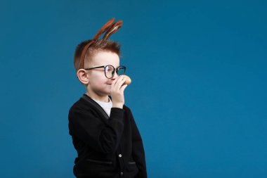 Mutlu Paskalyalar çocuklar. Tavşan kulaklı çocuk mavi arka planda renkli yumurta. Gözlüklü neşeli gülen çocuk..