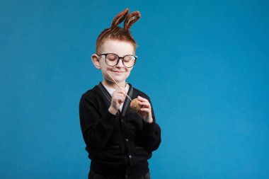 Mutlu Paskalyalar çocuklar. Tavşan kulaklı çocuk mavi arka planda fırçayla paskalya yumurtası boyuyor. Gözlüklü neşeli gülen çocuk..