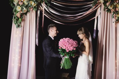 Damat, arka planda şık dekorasyon ve çiçeklerin, elektrik ampullerinin ve çelenklerin arkaplanına karşı büyük bir buket pembe gül sunar. Gece töreni.