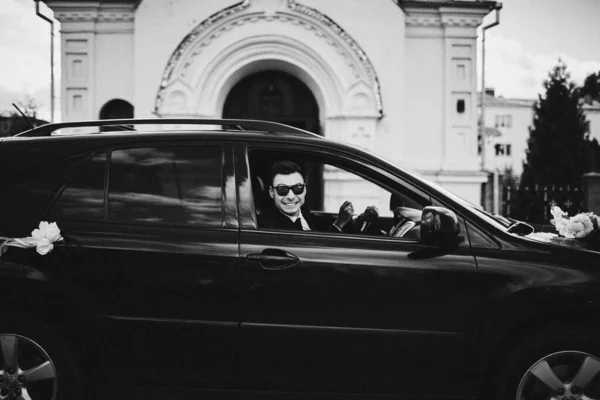 サングラスのハンサムな新郎は豪華な装飾車の中に座っている スーツ姿の男が結婚式の日に運転してる 白黒写真です — ストック写真