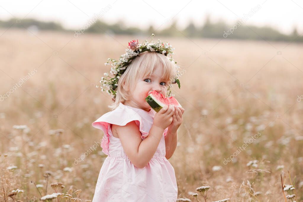 picnic with family. cute little girl eating big piece of watermelon on the grass in summertime in the park. Adorable child wearing in flowers wreath on head and pink dress