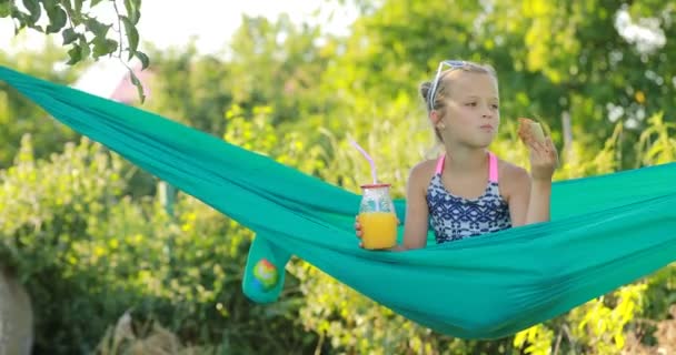 Portret szczęśliwego dziecka cute girl jest jedzenie pizzy i pić sok pomarańczowy ze słoika ze słomką siedzi na zielonym hamaku w ciepły dzień. letnie wakacje — Wideo stockowe