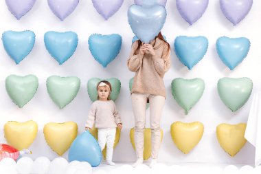 Güzel, genç bir anne ve küçük tatlı bir kız birlikte eğleniyorlar. Renkli kalp balonları üzerinde duruyorlar ve ellerinde mavi sevgililer günü balonu var. Doğum günü, Velentines, Anneler Günü