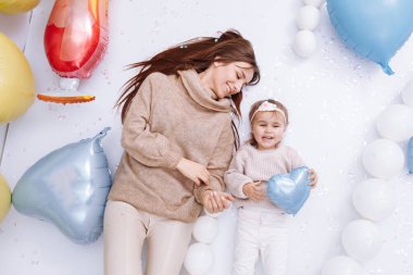 Güzel, genç bir anne ve küçük tatlı bir kız birlikte eğleniyorlar. Renkli balonların üzerinde duruyorlar. Arka planda, sarılıp öpüşüyorlar. Doğum günü, Velentines, Anneler Günü