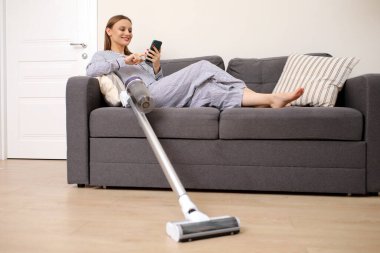 Kanepede oturan ve evi temizlerken cep telefonu kullanan genç bir kadın. Ev işleri ve ev işleri konsepti. Temizlik aletleri.