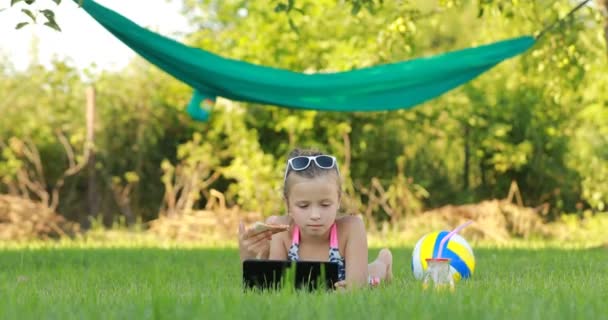 Dítě dívka je jíst kus pizzy a hrát na notebooku ležící na trávě v létě slunečný den. dítě obědvá po aktivních hrách. houpací síť a míč na pozadí.RIVNE, UKRAINE -červen 2021. — Stock video