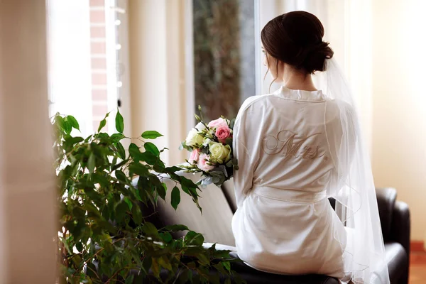 花嫁は白や淡いピンクのバラのブライダル花束を維持しており ウィンドウに座っています 花嫁の朝の集会だ バックビュー — ストック写真