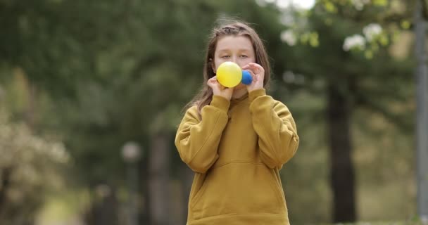 Portrait of cute adorable kid girl blow inflate blue and yellow air balloons for carrying Ukrainian day, green park background — Stock Video