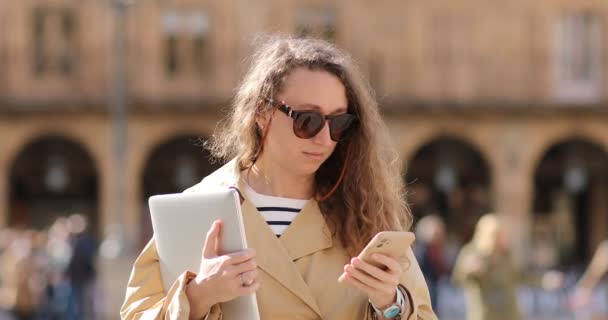 Uśmiechnięta młoda kobieta nosi strój relaks za pomocą telefonu komórkowego wpisując wiadomości tekstowe spaceru trzymając laptopa poza telefonem komórkowym biznesu miasta smartfon — Wideo stockowe
