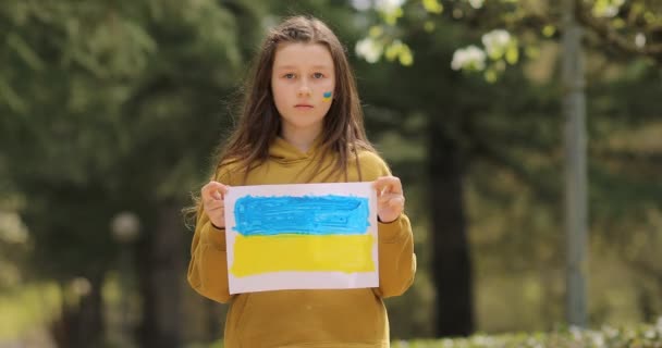 Cara seria de una niña. Las mejillas de los niños están pintadas con los colores amarillo y azul de la bandera de Ucrania. Los niños piden paz. — Vídeos de Stock