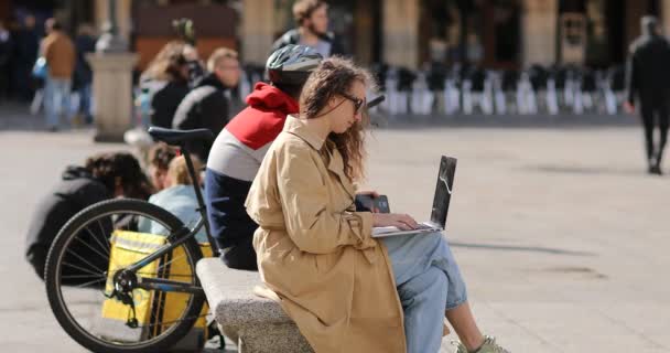 La jeune fille utilise un ordinateur portable assis à l'extérieur. Belle jeune femme bouclée avec ordinateur portable ayant un travail en ligne ou de l'éducation tout en étant assis sur les escaliers au milieu de la ville — Video