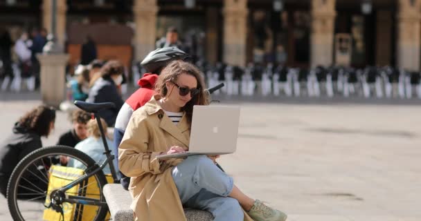 Salamanque, Espagne- Mars 2022 : Jeune fille utilise un ordinateur portable assis à l'extérieur. Belle jeune femme bouclée avec ordinateur portable ayant un travail en ligne ou de l'éducation tout en étant assis sur les escaliers au milieu de la ville — Video