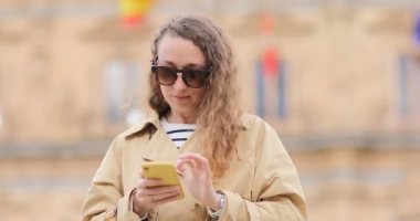 Şehir dışındaki caddede bir kadın cep telefonunu kullanıyor. Güneş gözlüklü neşeli turist eski İspanya şehrinin sokaklarında yürüyüp bir tanıdığa el sallıyor.