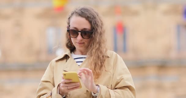 La mujer está usando el celular en la calle de la ciudad al aire libre. Alegre turista en gafas de sol divirtiéndose paseando por las calles de la vieja ciudad de España y saludando con la mano a un conocido — Vídeo de stock