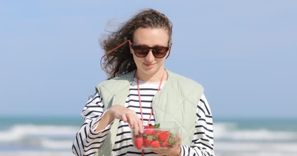 Χαμογελαστή νεαρή γυναίκα που απολαμβάνει φράουλες την ηλιόλουστη καλοκαιρινή μέρα, χαρούμενη ελκυστική γυναίκα με σγουρά μαλλιά που τρώει φρούτα κατά τη διάρκεια της ανάπαυσης μετά το περπάτημα κοντά στη θάλασσα — Αρχείο Βίντεο