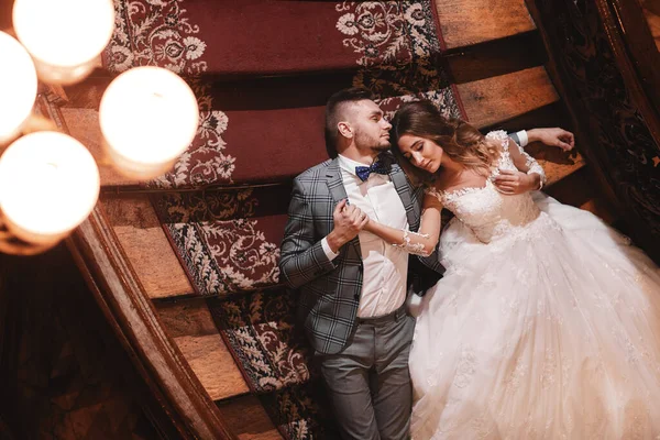 Ημέρα του γάμου. όμορφη νύφη σε λευκό μακρύ φόρεμα και νεαρός γαμπρός φορώντας μαύρο κοστούμι αγκαλιάζονται και βρίσκονται στις σκάλες μέσα σε μεγάλο ιστορικό κτίριο της Ευρώπης — Φωτογραφία Αρχείου