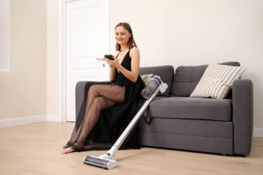 Uzun, zarif siyah elbisesi olan gülümseyen genç kadın koltukta oturuyor ve elinde elektrikli süpürge ile cep telefonu kullanıyor..