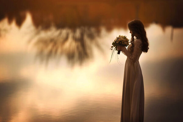 Bride in white dress with wedding bouquet near the lake at sunset