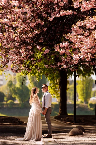 活泼的新婚夫妇 新郎和新娘拥抱着绽放的粉色樱花 一个笑容可亲的新郎和一个可爱的新娘的婚纱照 — 图库照片