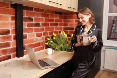 Çevrimiçi aşçılık dersi. Modern mutfakta elinde tencereyle blog yazarken dizüstü bilgisayar kullanan bir kadının kesilmiş fotoğrafı..