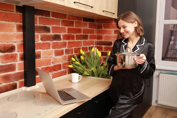 Çevrimiçi aşçılık dersi. Modern mutfakta elinde tencereyle blog yazarken dizüstü bilgisayar kullanan bir kadının kesilmiş fotoğrafı..